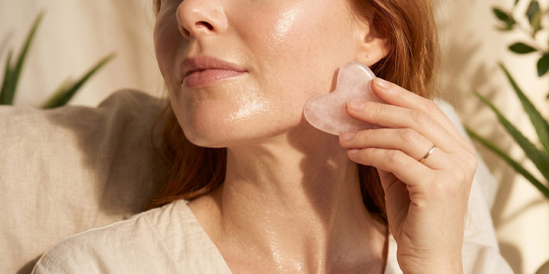 Woman using a pink gua sha tool on her face with a natural background.